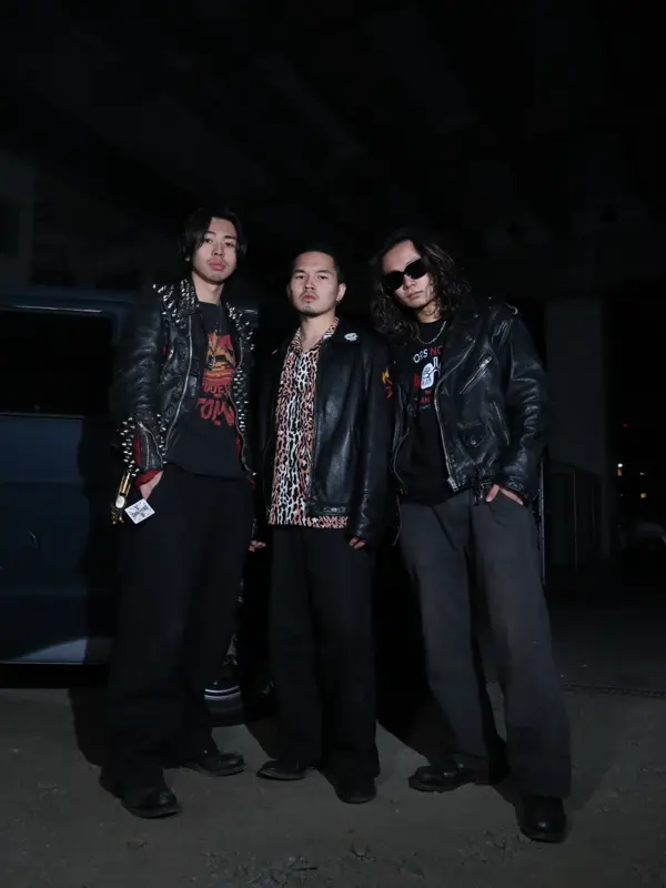 夜の屋外で並んで立つ、革ジャンを着たメンバー3人